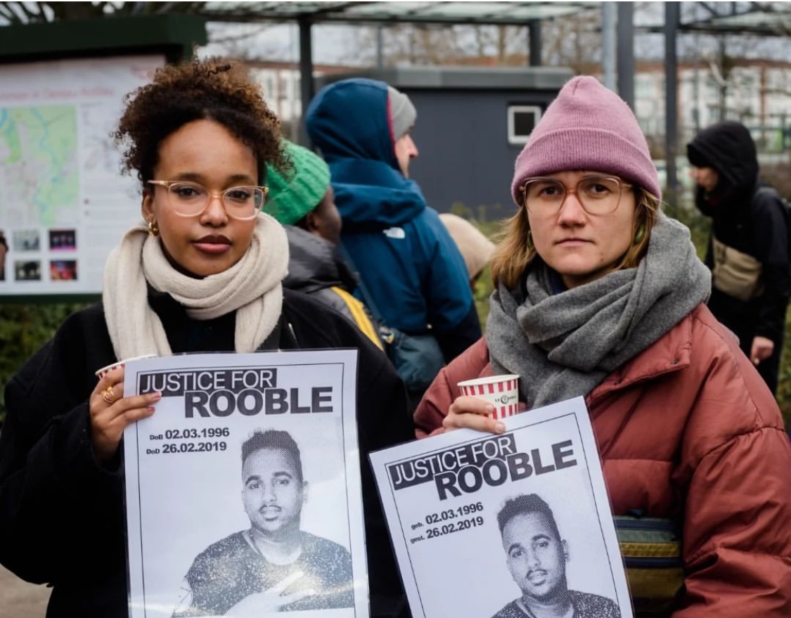 an image of two members of the justice 4 rooble campaign with somber faces holding posters that read 'justice for rooble, DOB 02.03.1996, date of death 26.02.2019'. 